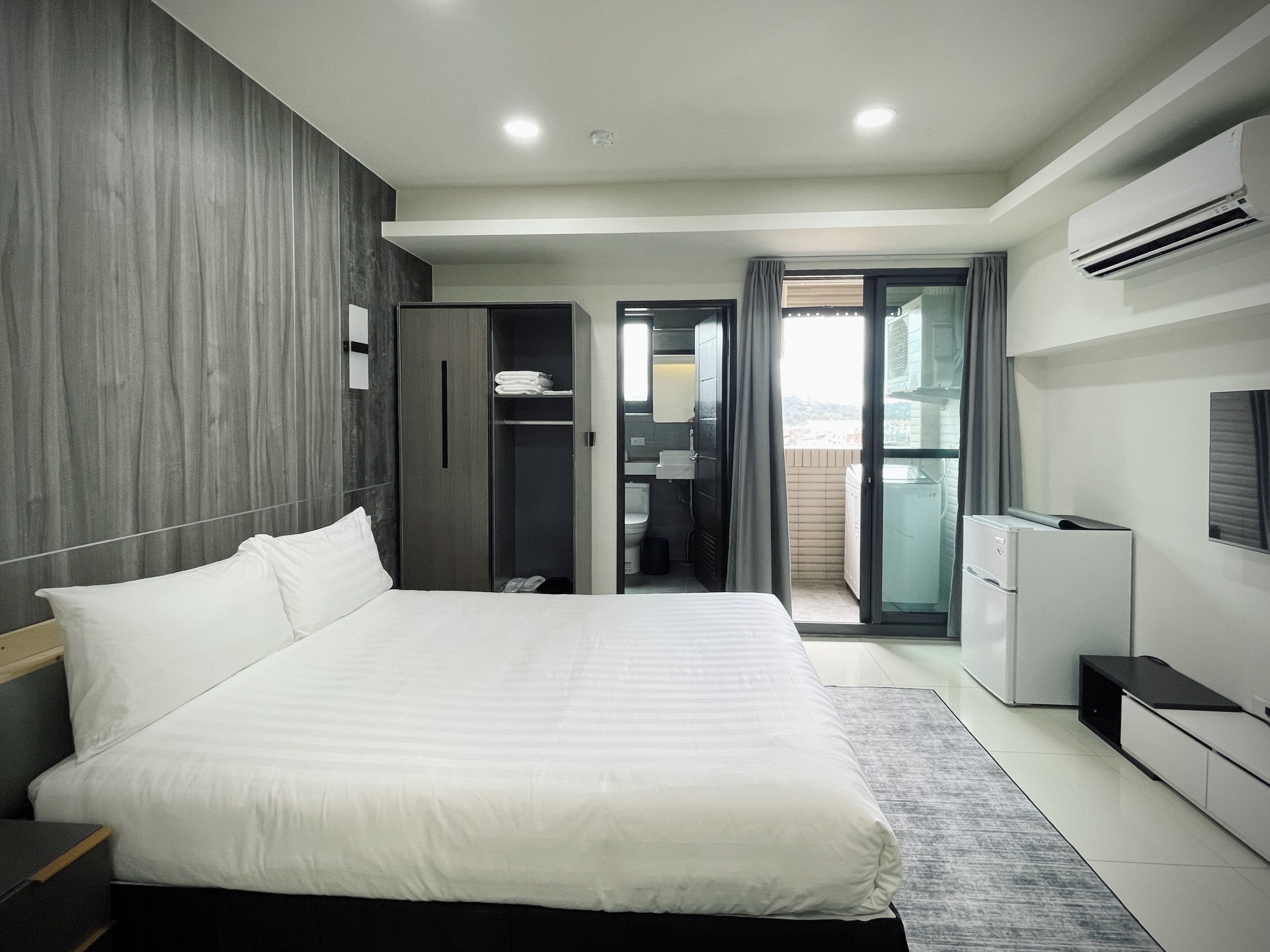 A modern hotel room with a large bed, a wardrobe, a bathroom with a toilet and sink, and a balcony with a view.