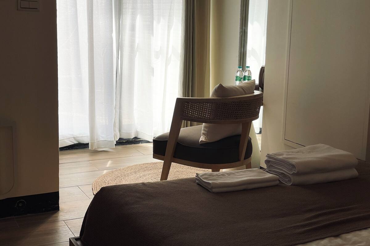 A hotel room with a bed, a chair, a window with curtains, and a bottle of water on the nightstand.