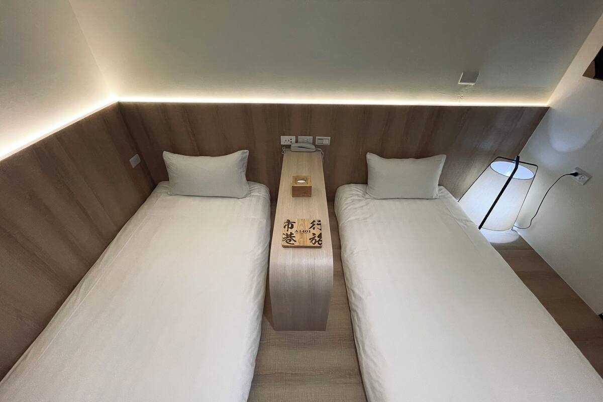 A hotel room with two single beds, a bedside table with a lamp, and a wall-mounted power outlet.