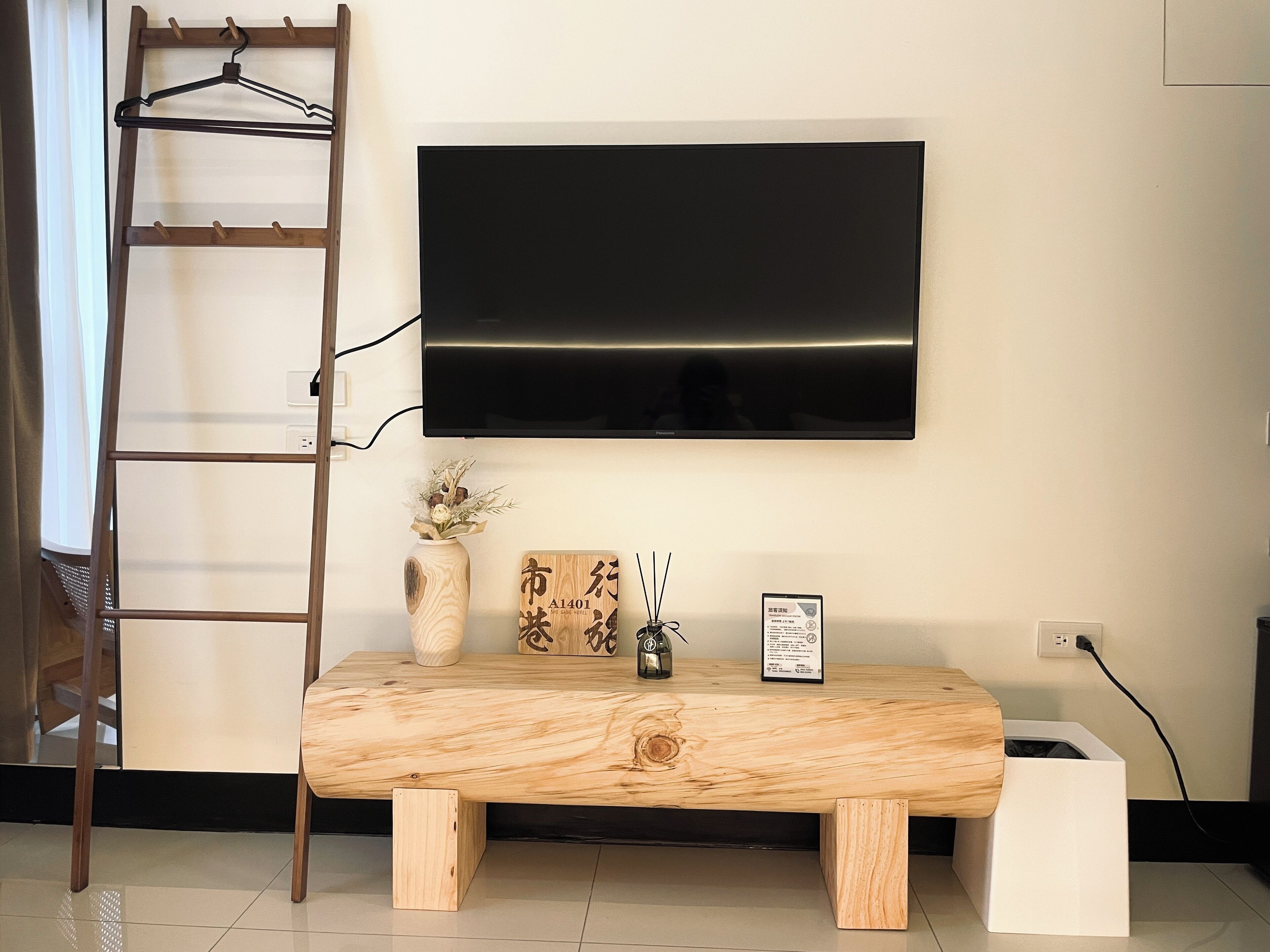 Un salon moderne avec un banc en bois, un téléviseur fixé au mur, un petit vase contenant des fleurs séchées et une échelle appuyée contre le mur.
