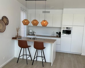 Cozinha moderna com armários brancos, ilha de madeira com dois banquetas e luminárias pendentes.