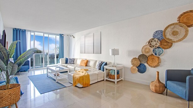 Uma sala de estar moderna com um sofá branco, uma poltrona azul e uma mesa de centro. Há cestos decorativos de fibra na parede e uma grande planta em vaso.