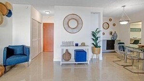 Uma sala de estar moderna com um sofá azul, uma mesa consola branca com objetos decorativos, uma área de jantar com mesa de vidro e cadeiras de metal, e uma cozinha com fogão e armários.