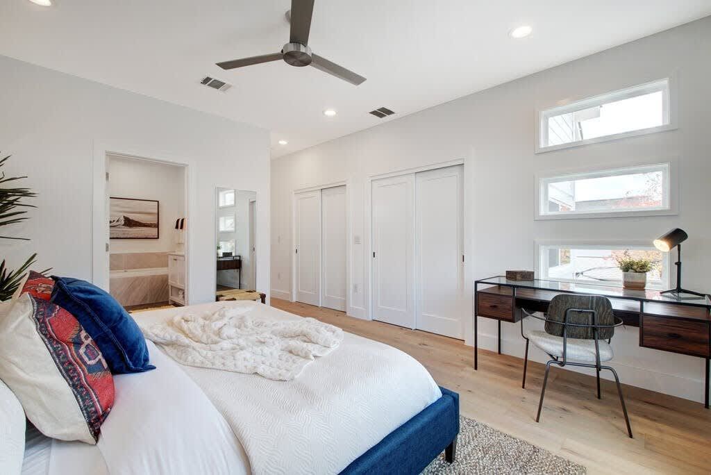 A bedroom with a bed, a desk, and a chair. There is a ceiling fan, a window, and a door.