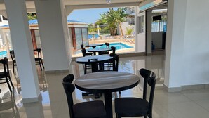 Zona de piscina con mesa redonda y sillas, suelo de baldosas y vistas a una piscina y palmeras.