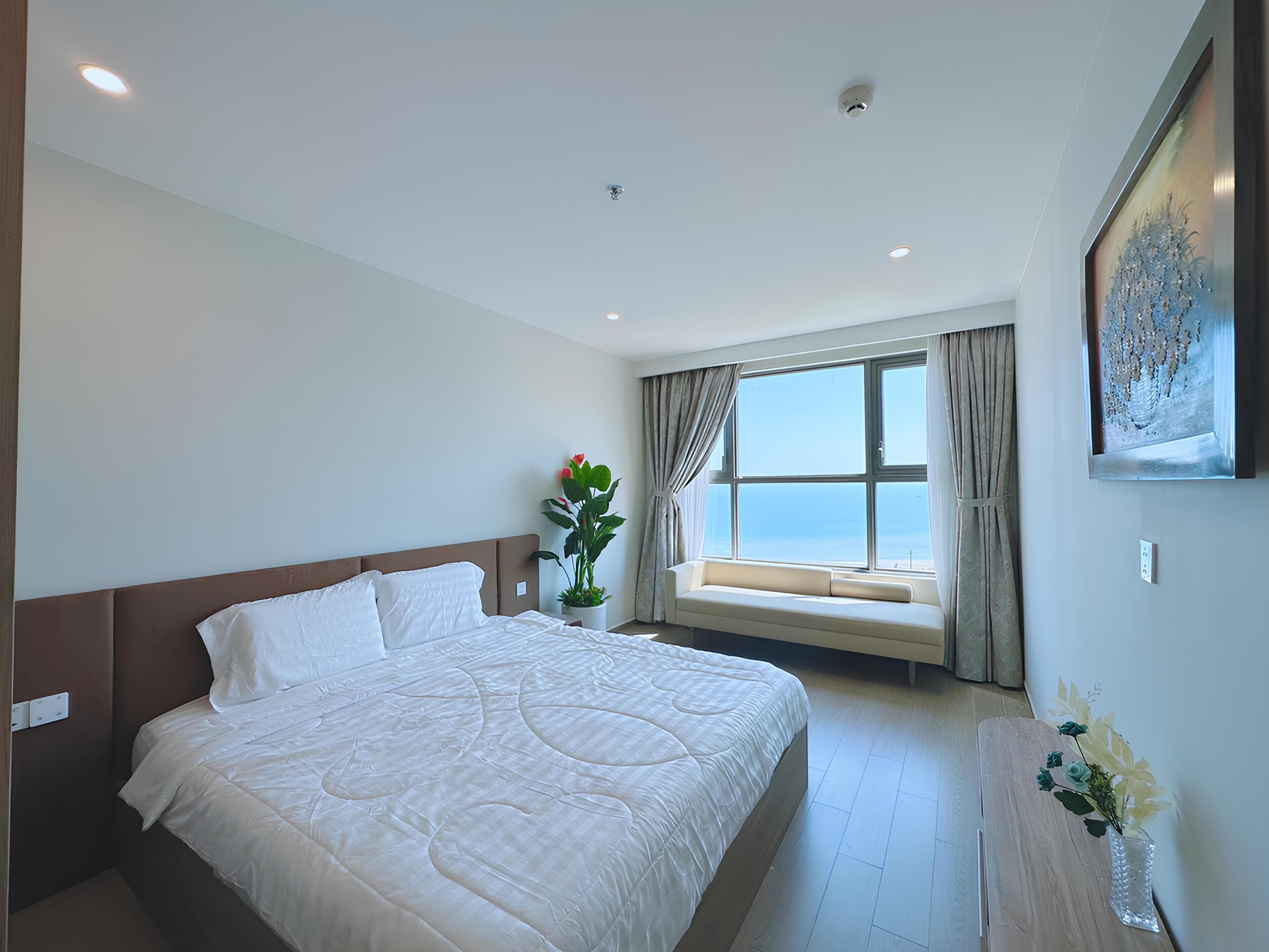 A hotel room with a large bed, a potted plant, a window with a view, and a painting on the wall.