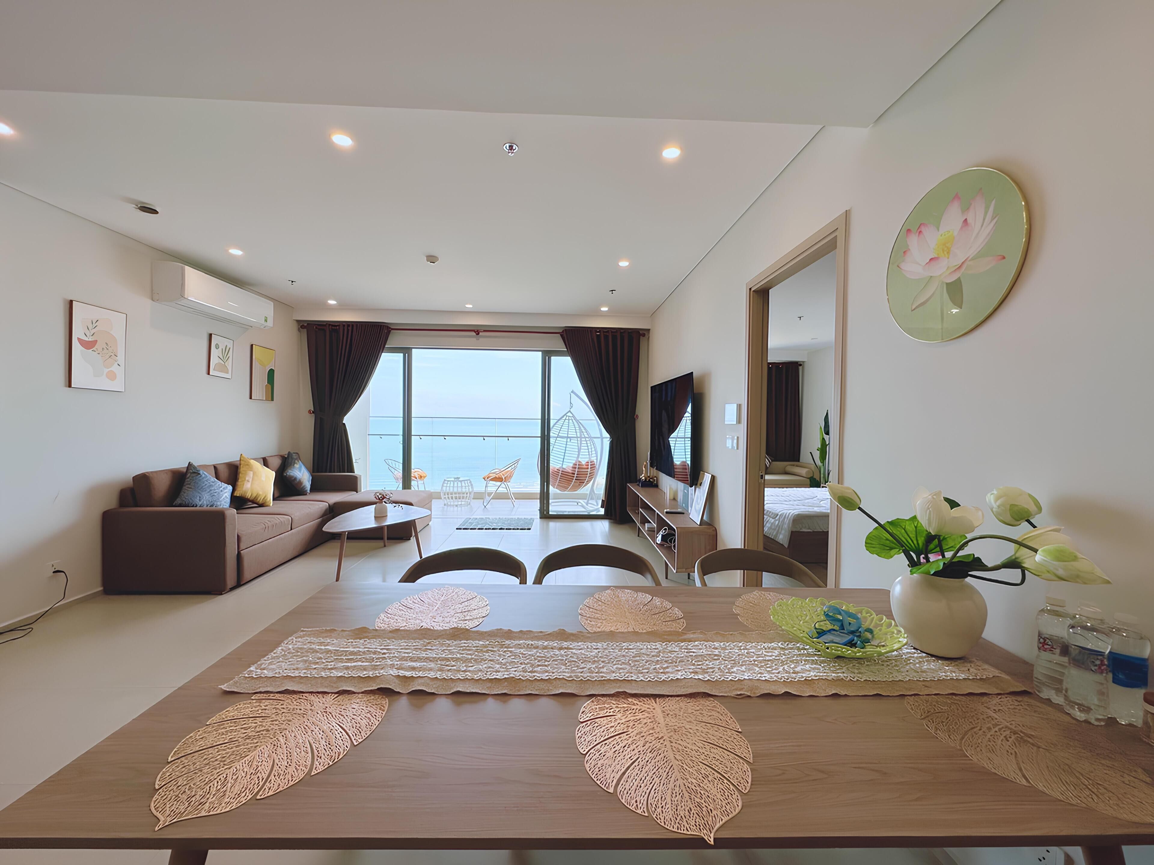 A modern living room with a large dining table, a sofa, and a view of the sea.
