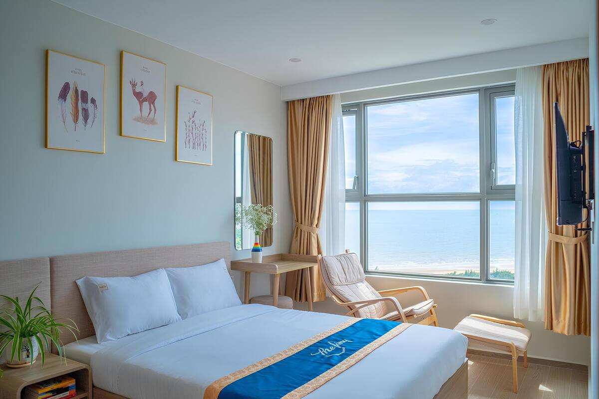 A hotel room with a bed, a desk, a chair, a TV, and a large window with a view of the sea.
