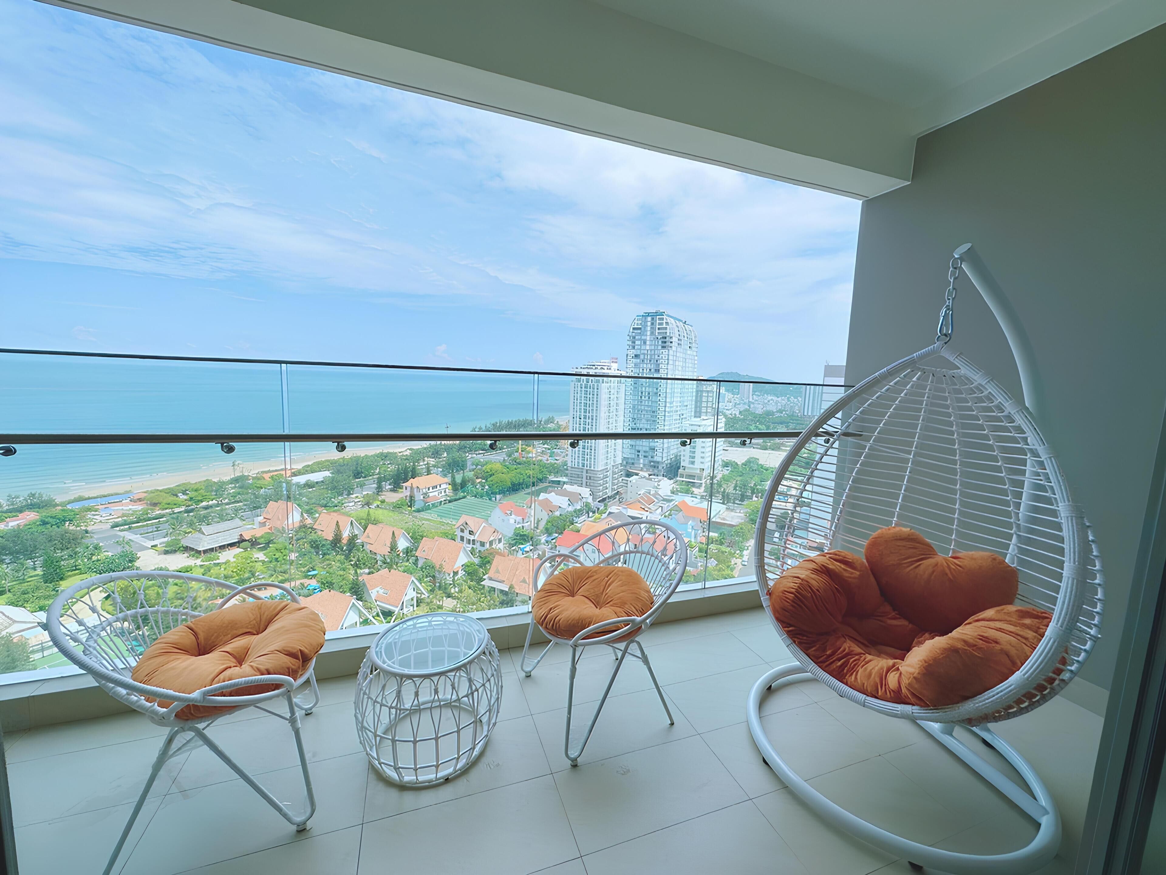 A balcony with a hanging chair and two chairs with cushions, overlooking a cityscape and beach.