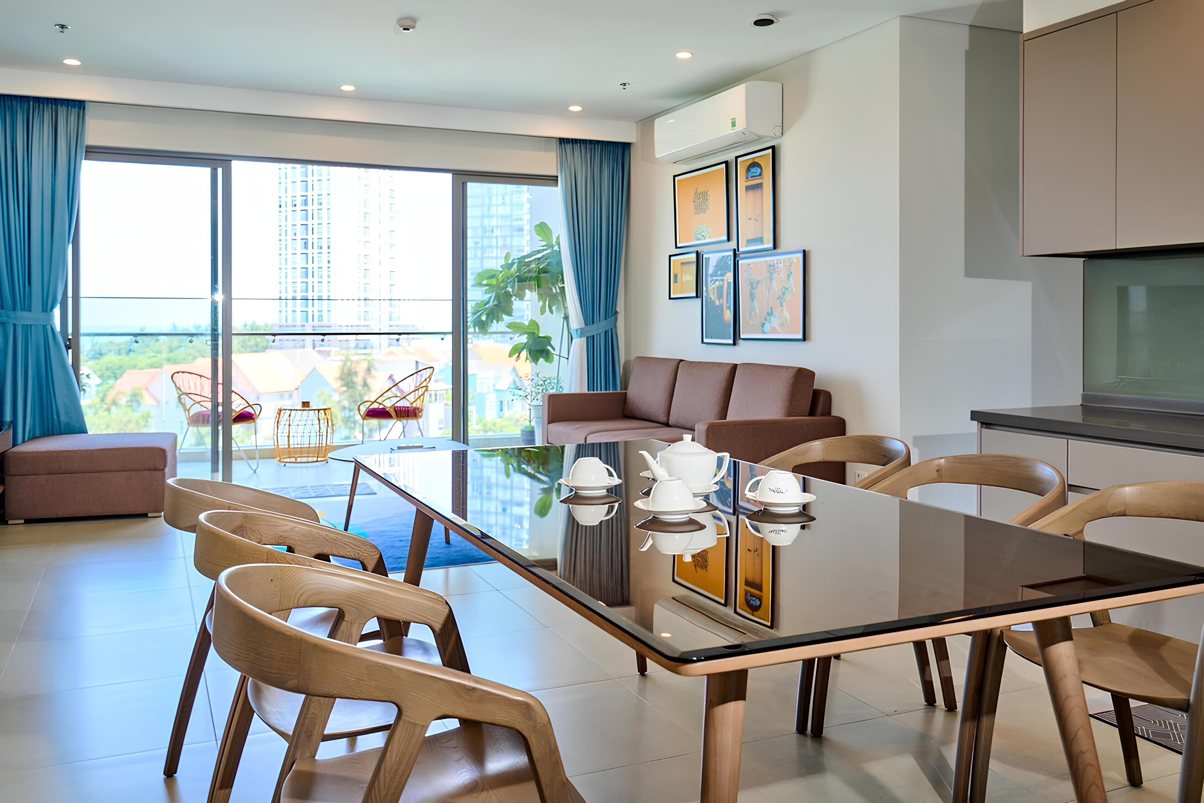 Una sala moderna con una mesa de comedor de cristal, sillas de madera, un sofá y un balcón con vistas a edificios y vegetación.