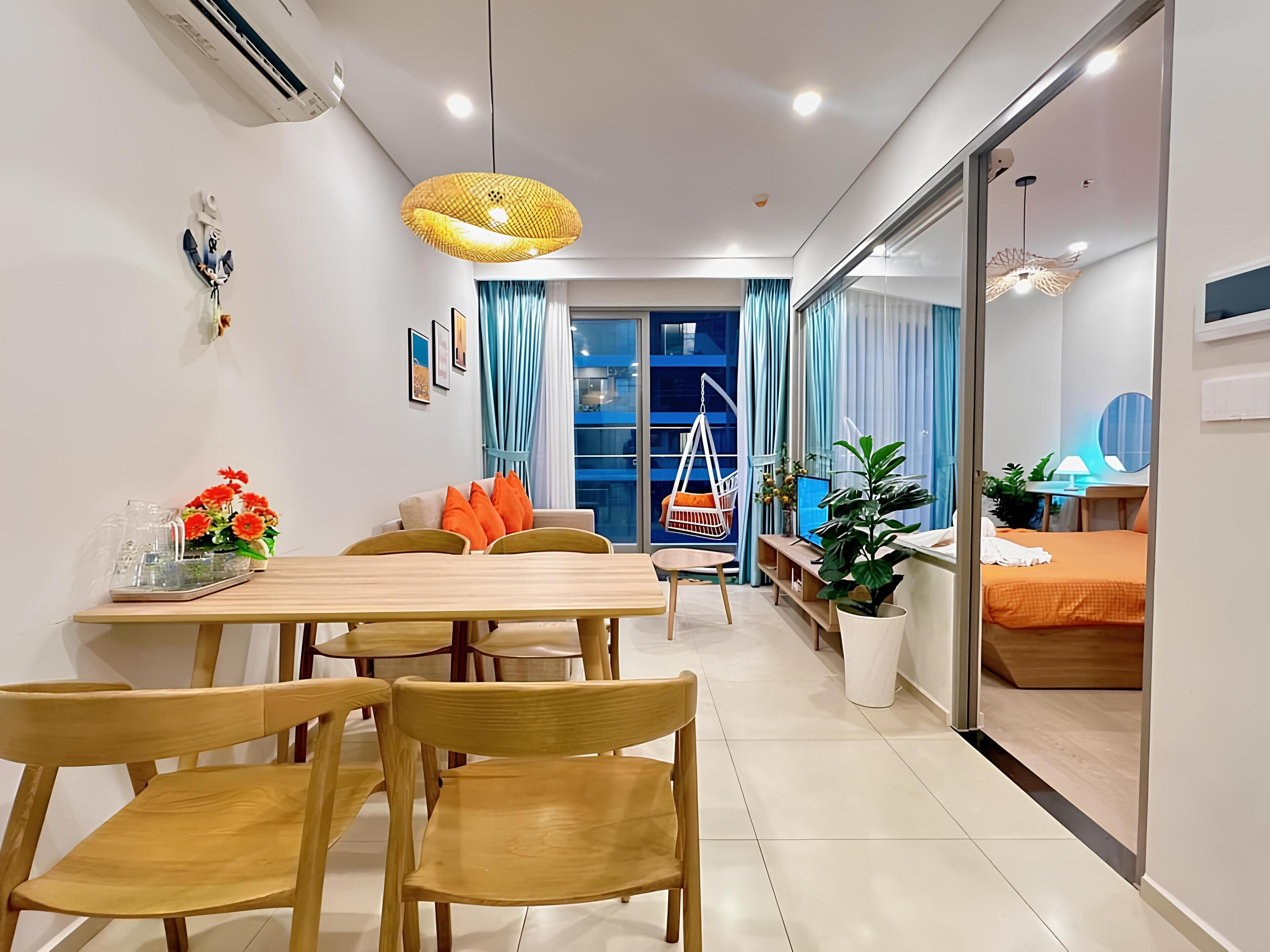 Une chambre d’hôtel moderne avec un coin repas, un lit recouvert d’une couverture orange, un bureau avec une lampe et une grande fenêtre ornée de rideaux bleus.