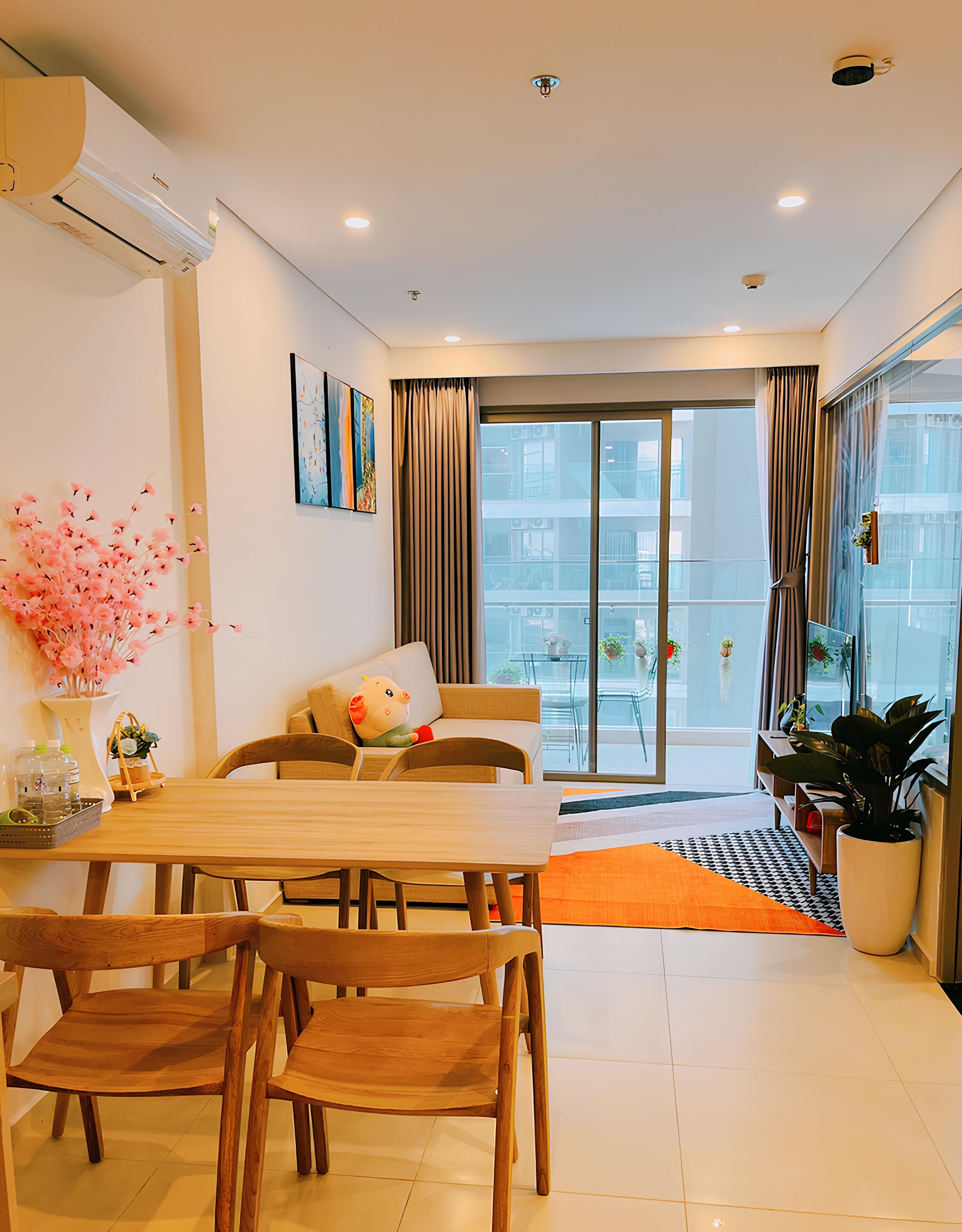 A modern living room with a wooden dining table, comfortable seating, a large window with curtains, and a balcony with plants.