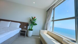 Una habitación de hotel moderna con una cama grande, una mesita de noche pequeña, una planta en maceta, un sofá junto a la ventana y vistas al mar.