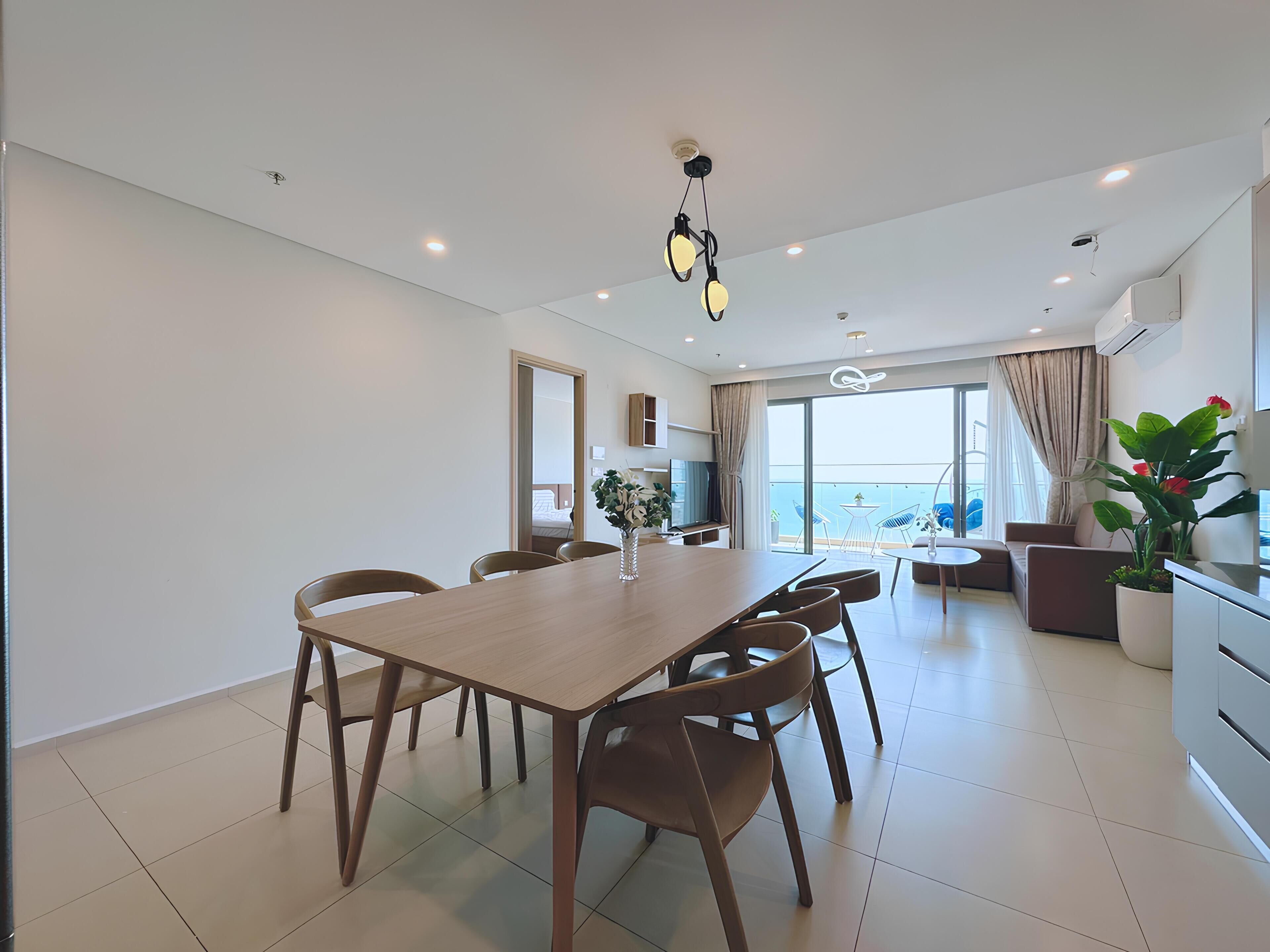 A modern living room with a wooden dining table, chairs, a sofa, and a balcony with a view.