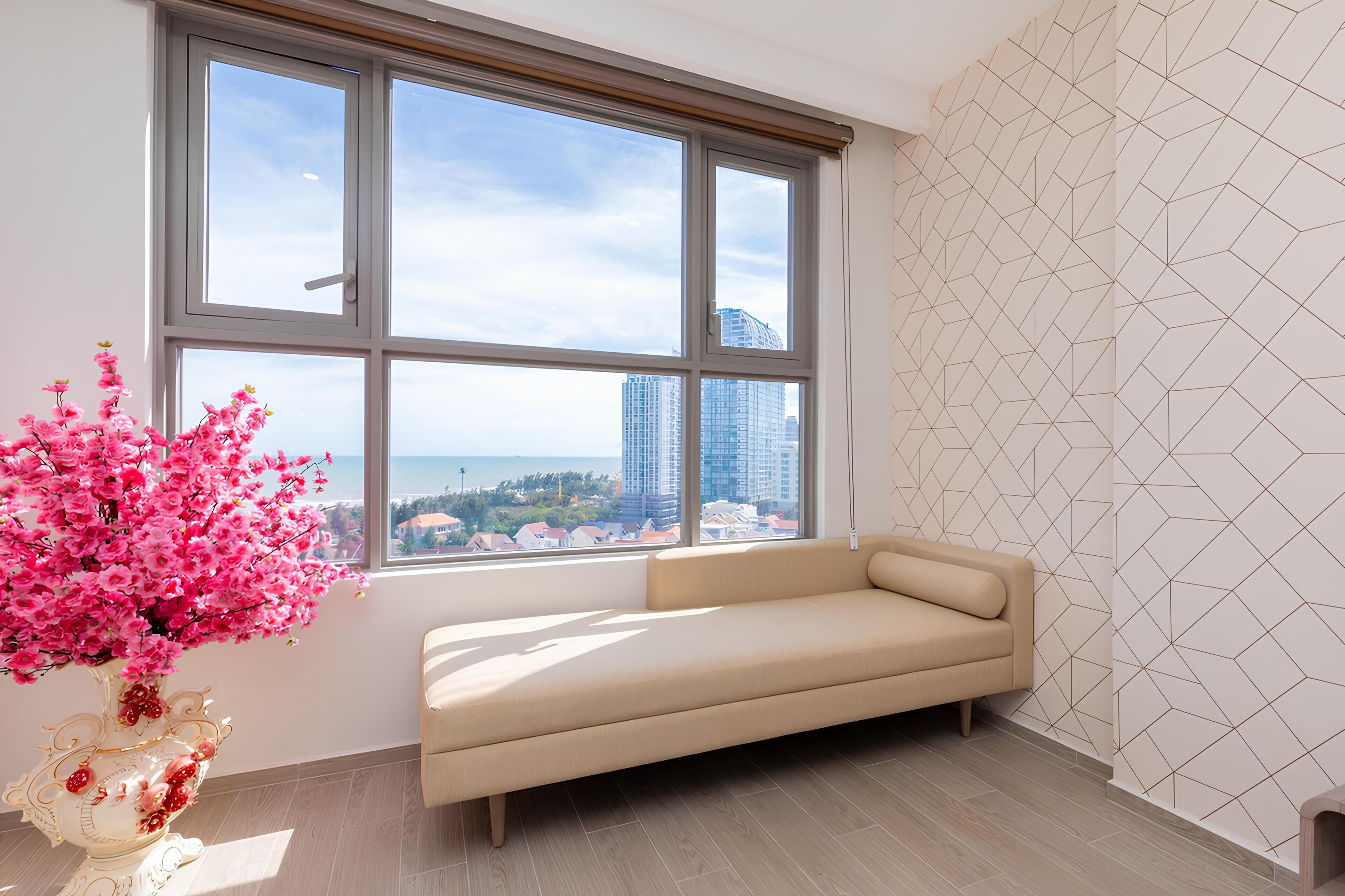 Um quarto com uma janela grande, um sofá bege e um vaso decorativo com flores cor-de-rosa.