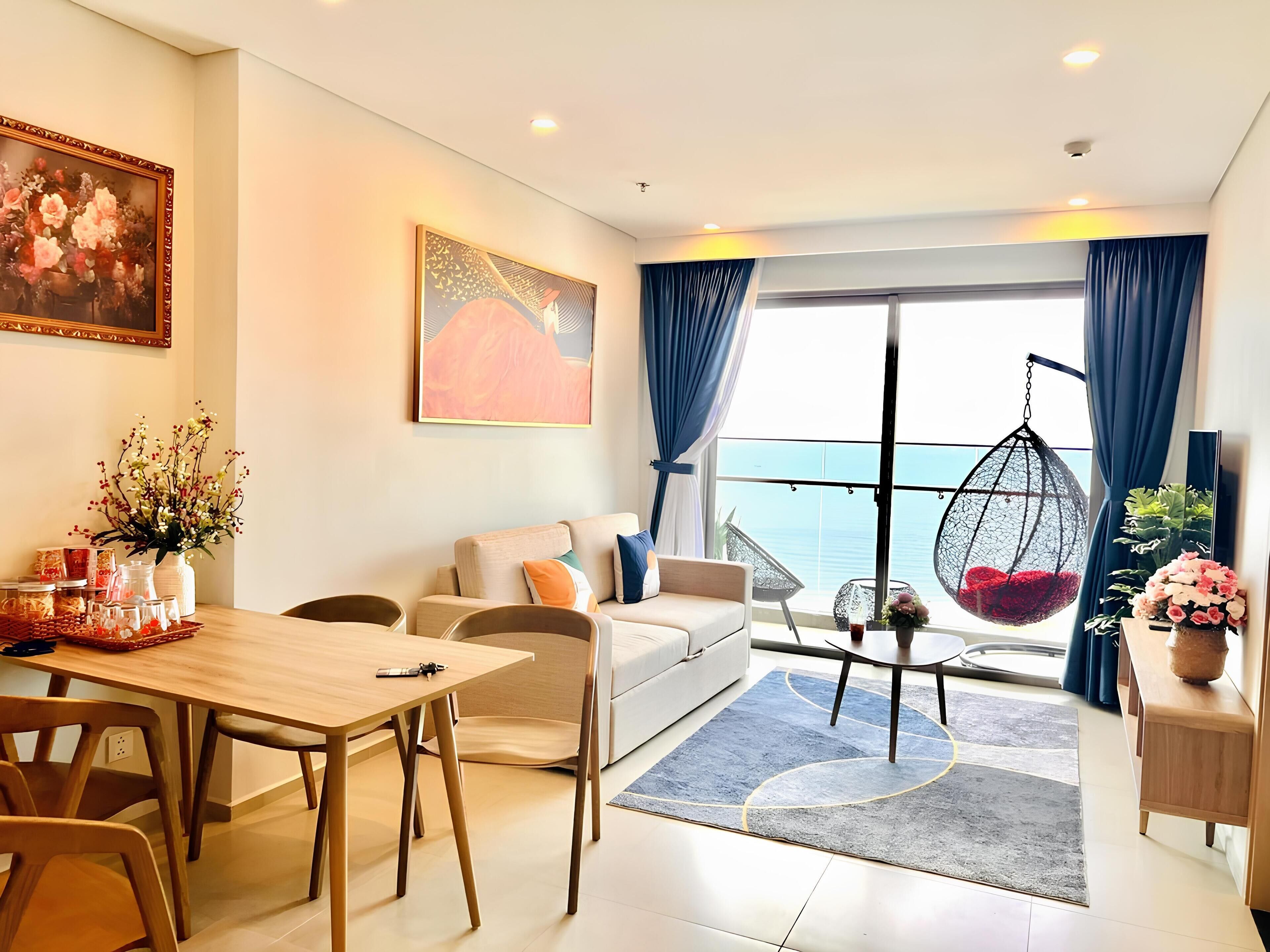 Un salon moderne comprenant un canapé, une table à manger et des chaises, un fauteuil suspendu, et offrant une vue sur la mer grâce à de grandes fenêtres.