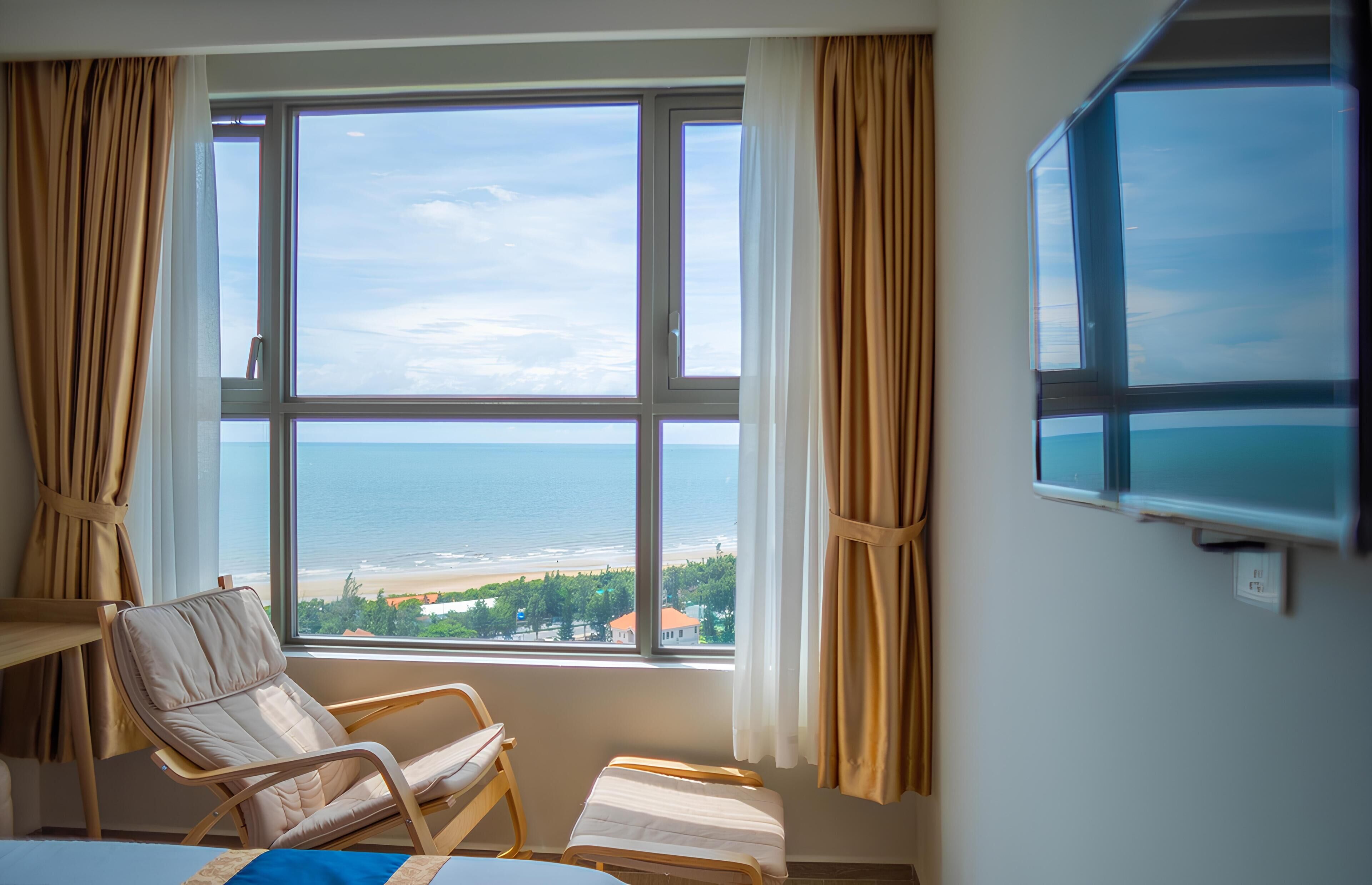 A room with a large window offering a view of the beach and ocean. There is a chair and a small table.