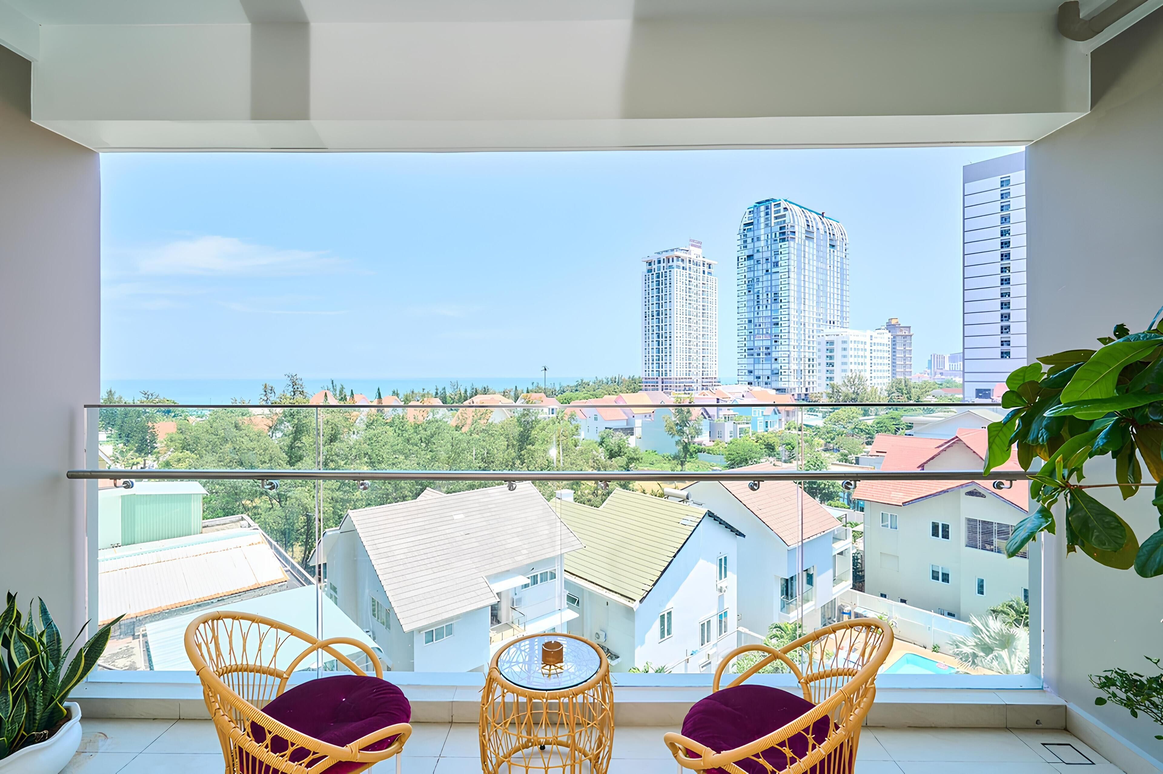A balcony with wicker chairs and a table, overlooking a cityscape with buildings and a clear sky.
