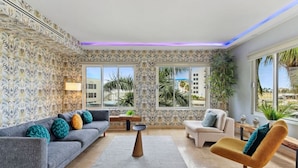 A modern living room with patterned wallpaper, a grey sofa with colorful cushions, a round coffee table, a beige armchair, and a view of palm trees and buildings through large windows. - Stunning 2BR/3Beds Lux Condo w/ Private Pool at Sunset 13 by Bocobay (Noord)