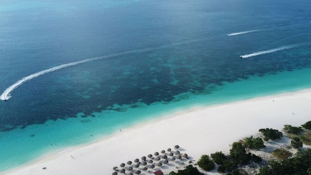 An aerial view of a beach with clear turquoise waters, a white sandy shore, and a row of round tables and umbrellas. - Stunning 2BR/3Beds Lux Condo w/ Private Pool at Sunset 13 by Bocobay (Noord)