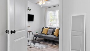 A modern living room with a grey sofa, a ceiling fan, a mounted TV, and a window with blinds. - Charming, Private Abode: Serenity in Hopewell, VA! (Hopewell)