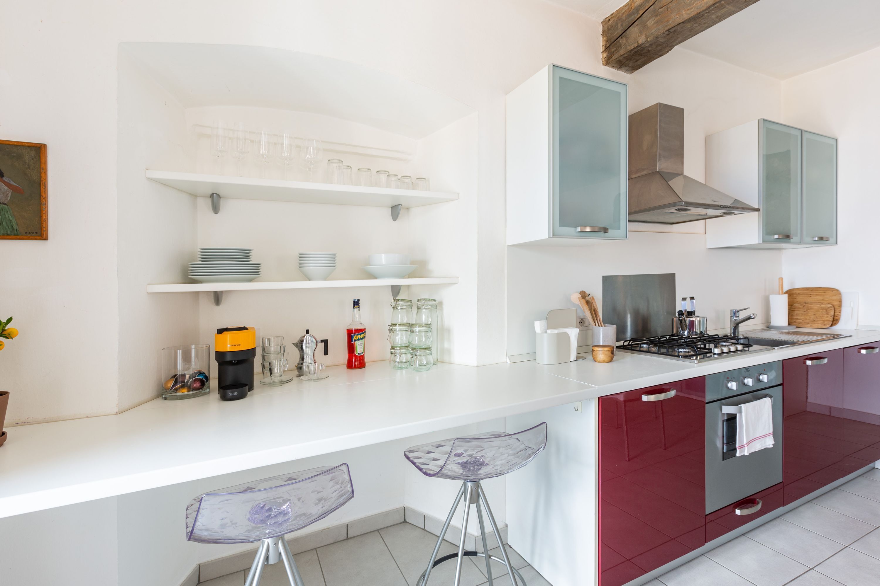 Una cocina moderna con encimeras blancas, armarios rojos y horno integrado. Hay taburetes de bar y una cafetera en la encimera.