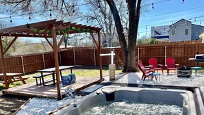 A backyard with a hot tub, wooden pergola, picnic tables, and red chairs.