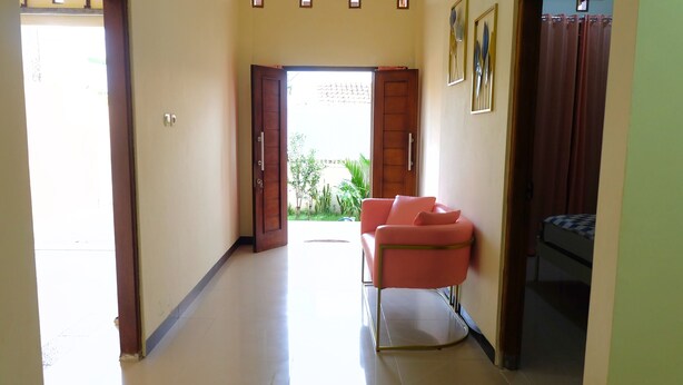 A hallway with a pink sofa, a bed with a checkered blanket, and a view of a garden through an open door. - Alesya Homestay Syariah (Yogyakarta)