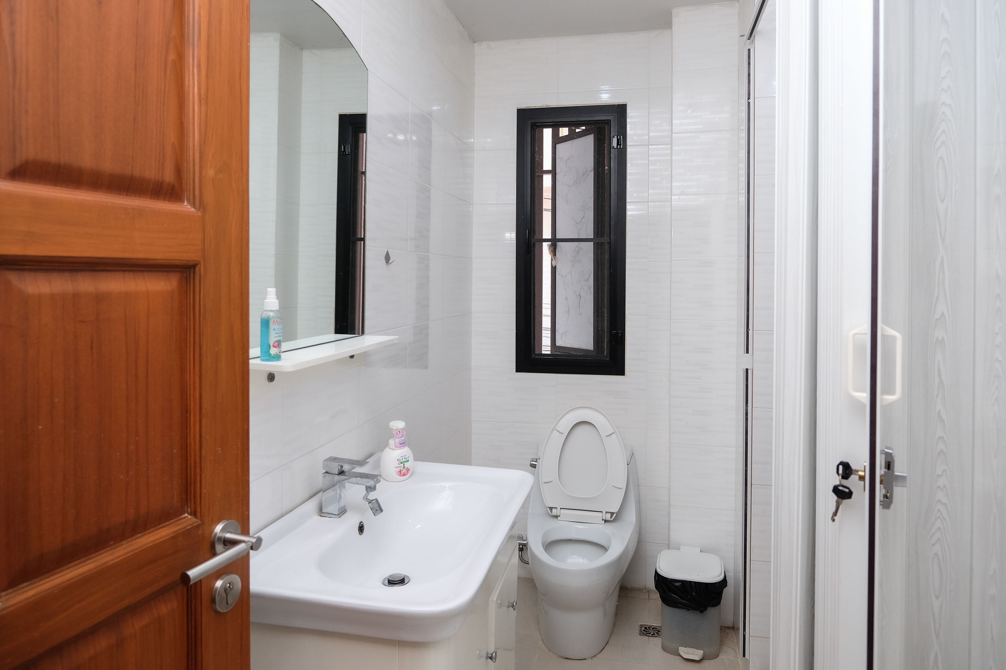 Un baño con puerta de madera, azulejos blancos, lavamanos blanco, inodoro y un cubo de basura.