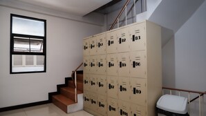 A stairwell with beige lockers labeled B1 to B12, a white trash bin, and a window with blinds. - Delta 9 Hostel (Bangkok)