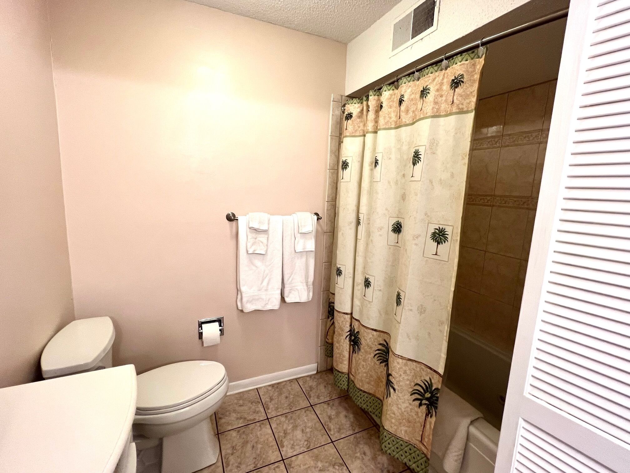 A bathroom with a toilet, a shower curtain with palm tree patterns, and a towel rack with two towels.