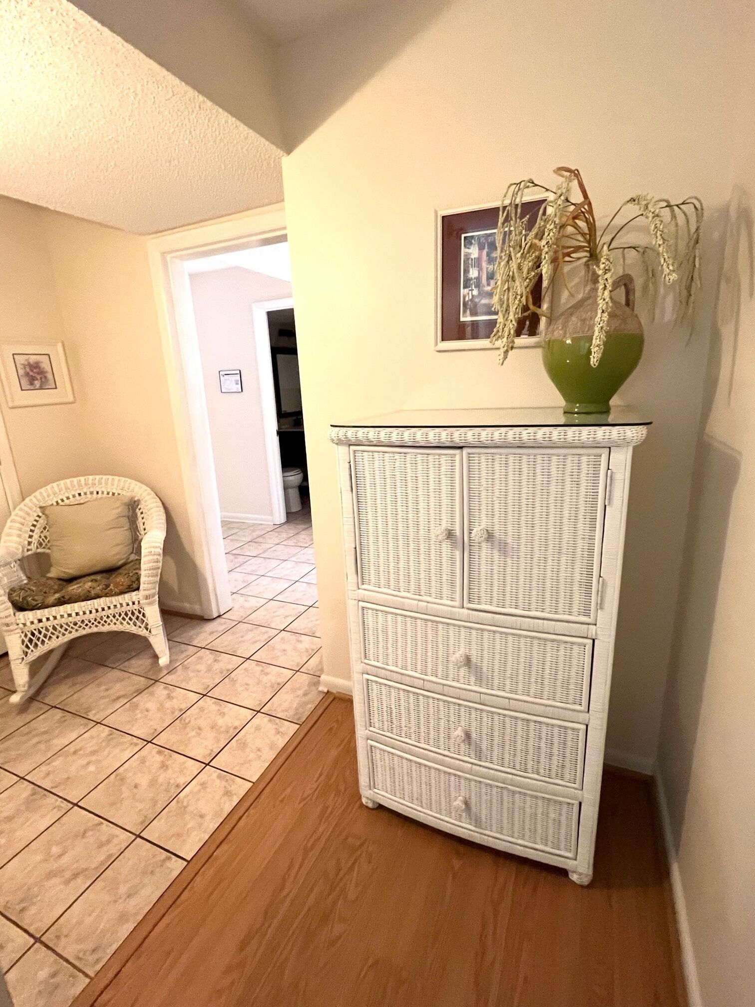 Une pièce comprenant un fauteuil en osier, une commode blanche et un vase vert contenant des plantes séchées.