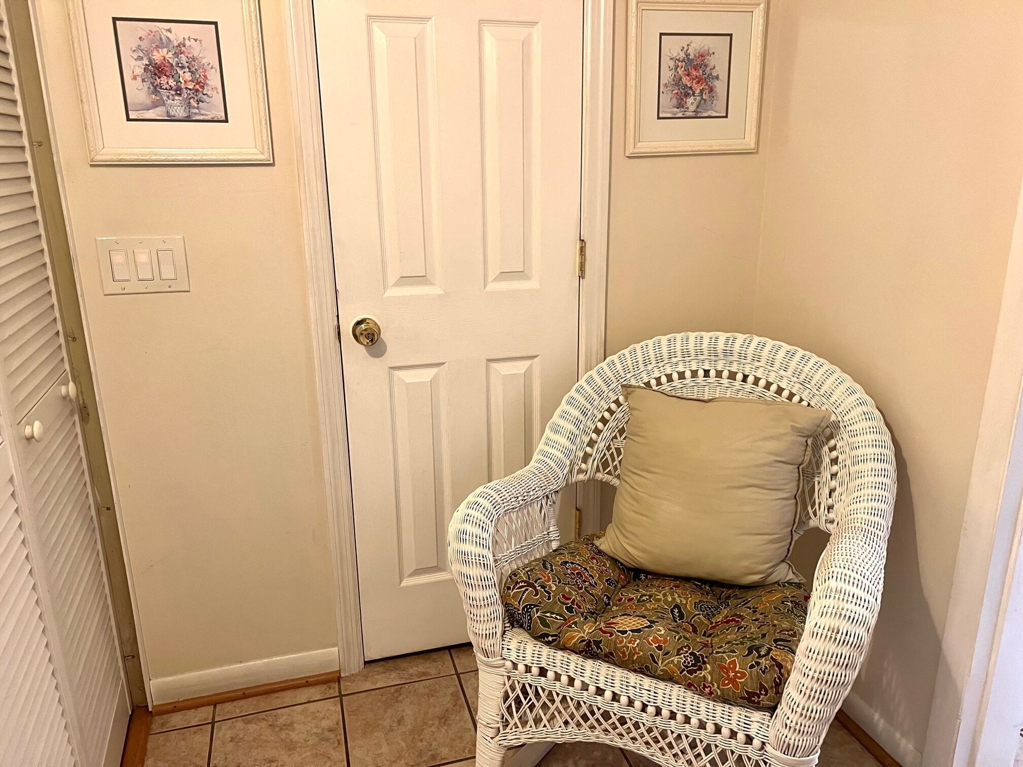 Une chaise en osier blanche, dotée d’un coussin à motifs, se trouve dans un couloir avec une porte blanche et deux tableaux floraux encadrés accrochés au mur.