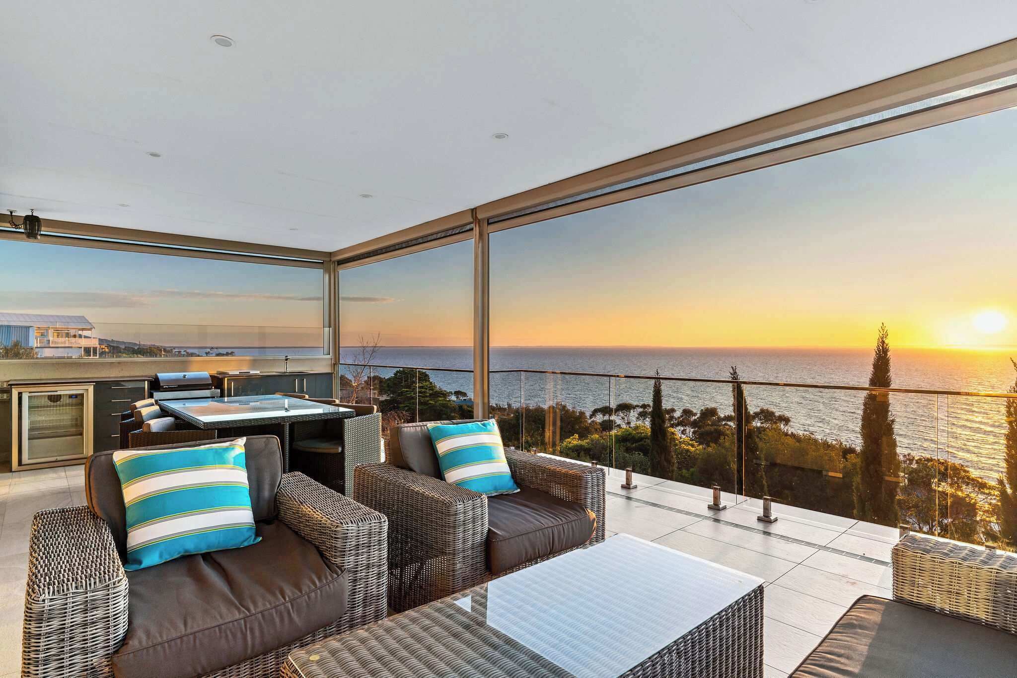 Un balcon agrémenté de meubles en osier, d’une table en verre et offrant une vue sur le coucher de soleil sur la mer.