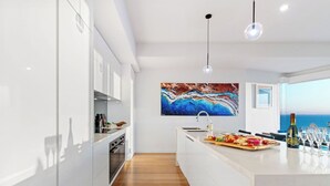 Uma cozinha moderna com armários brancos, piso de madeira, uma ilha central com um prato de frutas e uma grande pintura abstrata acima da pia.