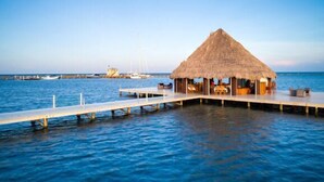 Een rieten dak bungalow op het water met een houten terras dat uitloopt in de zee, een gebouw op de achtergrond en boten afgemeerd aan een steiger.