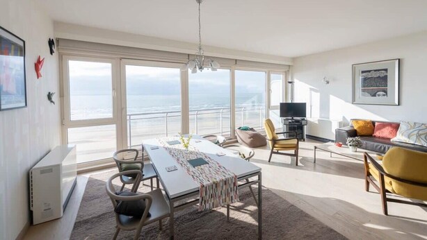 Un amplio salón con un gran ventanal con vistas al mar, una mesa de comedor con sillas, un sofá y un televisor.