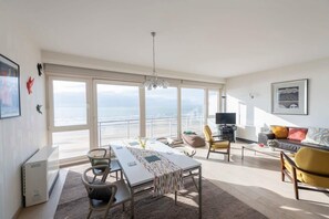 A spacious living area with a large window overlooking the sea, a dining table with chairs, a sofa, and a television. - Seaview Apartment on the Beach - Free Parking! (Oostende)