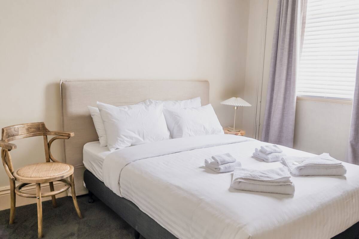 Een net opgemaakt bed met witte lakens, een houten stoel met een geweven zitting en een nachtkastje met een lamp.