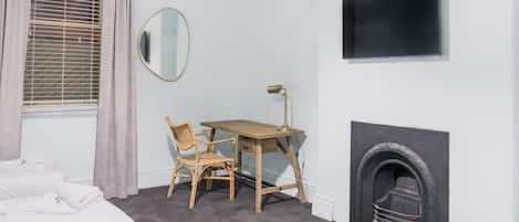 Un dormitorio con cama, escritorio y silla, espejo, chimenea y televisor en la pared.
