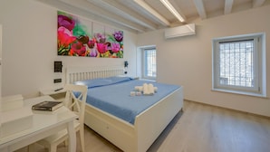 Un dormitorio con cama, escritorio, silla, cuadro, ventana y aire acondicionado.