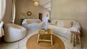 Un vaste espace de vie comprenant un canapé, un fauteuil et une table basse. Deux lampes suspendues et un miroir ornent le mur.