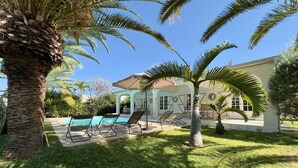 Un espace piscine avec des chaises longues, un patio couvert et une maison à l’extérieur de couleur claire.