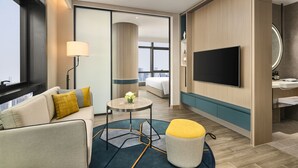 Ein modernes Hotelzimmer mit einer Sitzecke, einem runden Tisch, einem Pouf, einem Fernseher und einem Badezimmer mit Waschbecken und Spiegel.