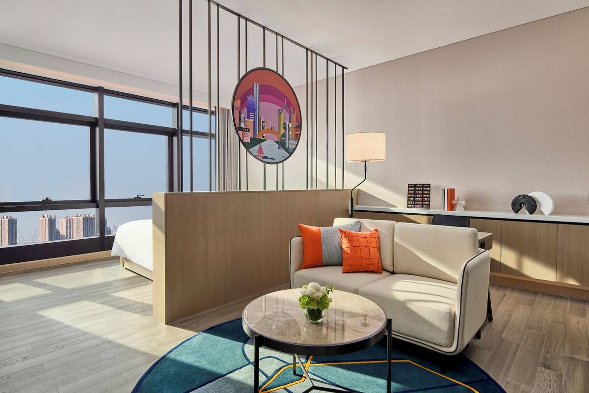 Een moderne hotelkamer met een groot raam, een ronde salontafel, een bank met kussens, een wandtelevisie en een bureau met een lamp.