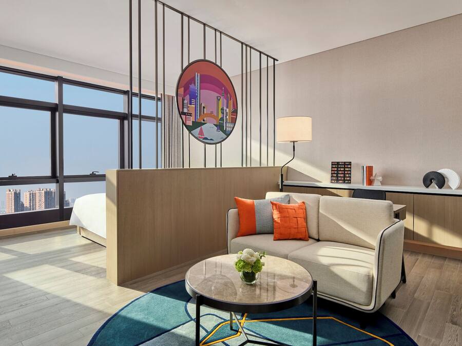 A modern hotel room with a large window, a round coffee table, a sofa with cushions, a wall-mounted TV, and a desk with a lamp.