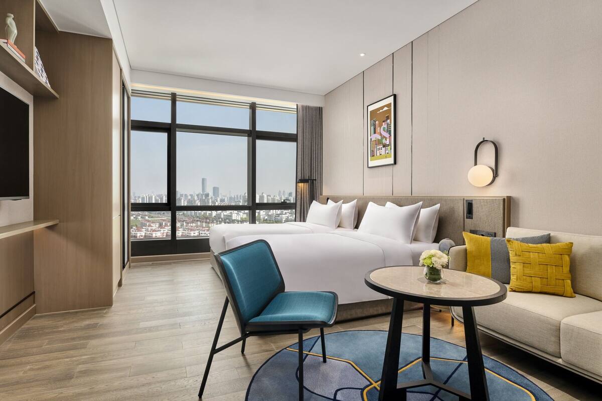 Een moderne hotelkamer met een groot bed, een slaapbank, een fauteuil, een klein tafeltje en uitzicht op de stad door grote ramen.