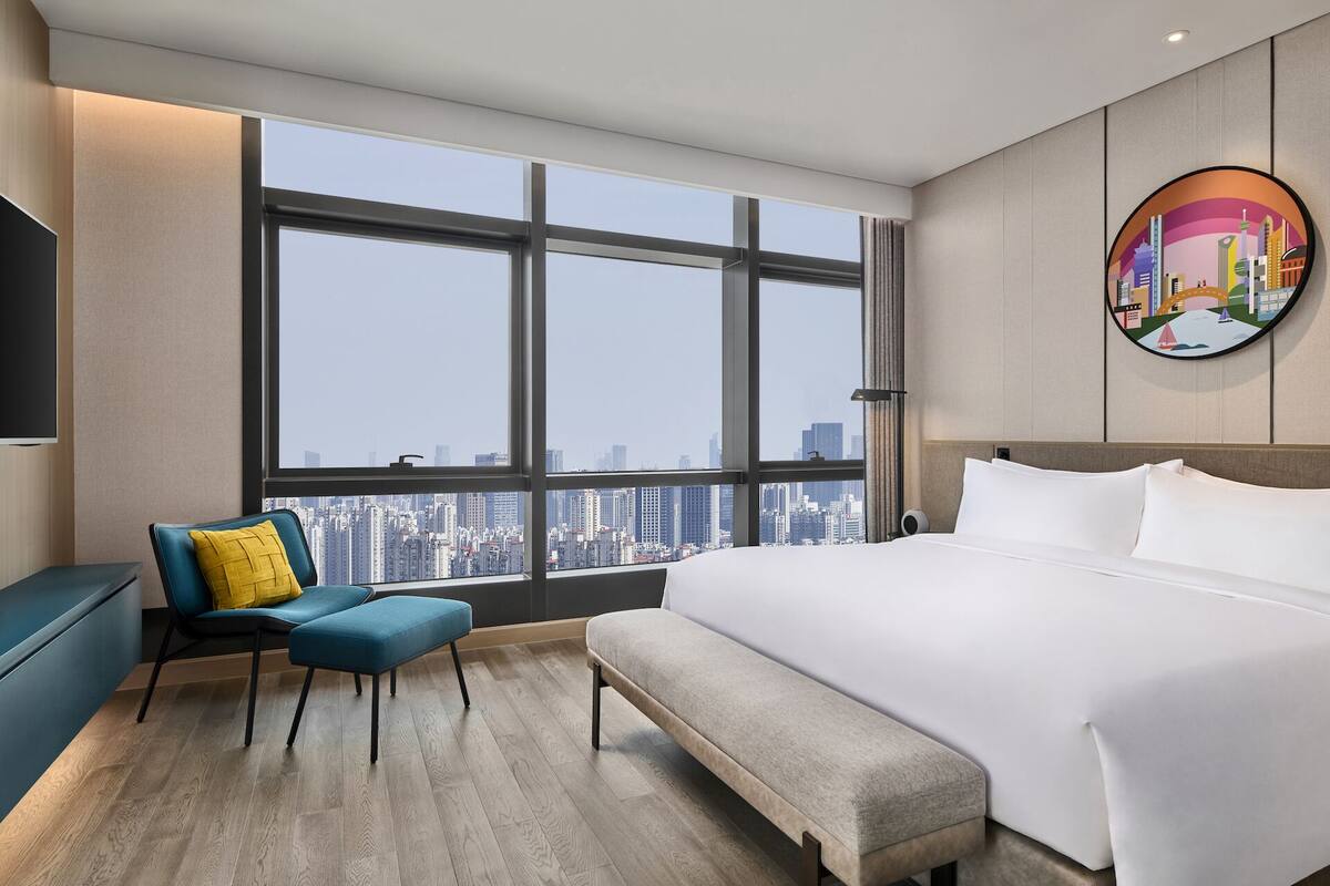 Een moderne hotelkamer met een groot raam, een bed, een bankje, een stoel en een televisie.