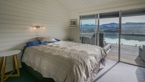 Ein Schlafzimmer mit einem großen Bett, einem kleinen Tisch und Blick auf einen See und Berge durch eine Glastür.
