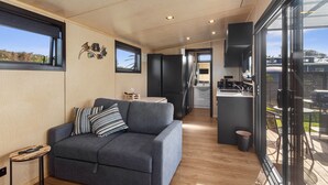 A modern living room with a grey sofa, wooden flooring, and a kitchen area visible through an open door. - Peace in Paradise - Tokerau Beach Holiday Home (Tokerau Beach)